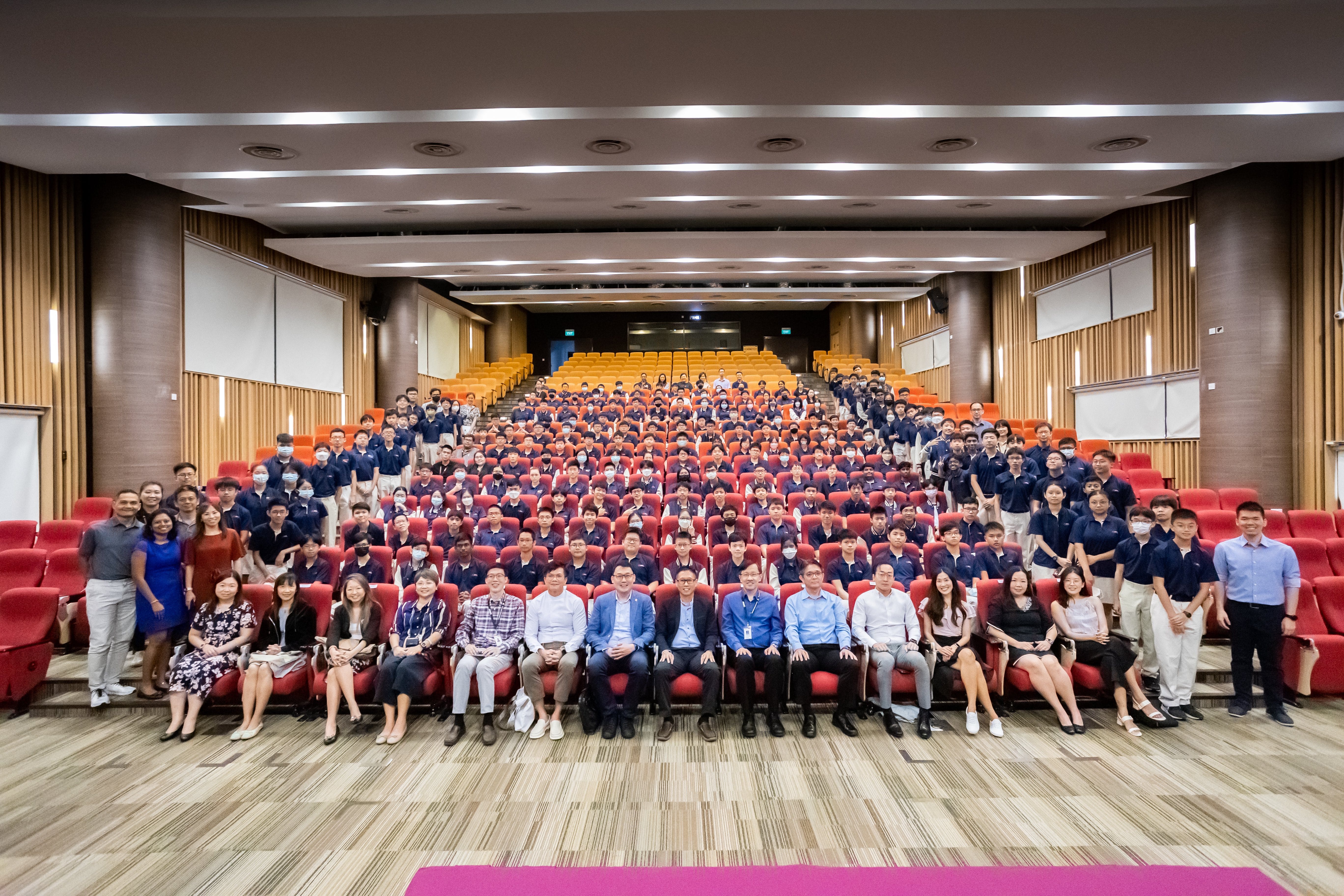 Large group photo in an auditorium with rows of red seats and many people posing, some wearing masks.